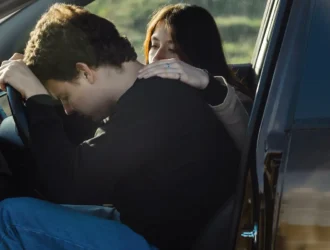 homem vestido de preto sentado em um carro com a cabeça encostada no volante com medo de dirigir e uma mulher ao seu lado o confrota