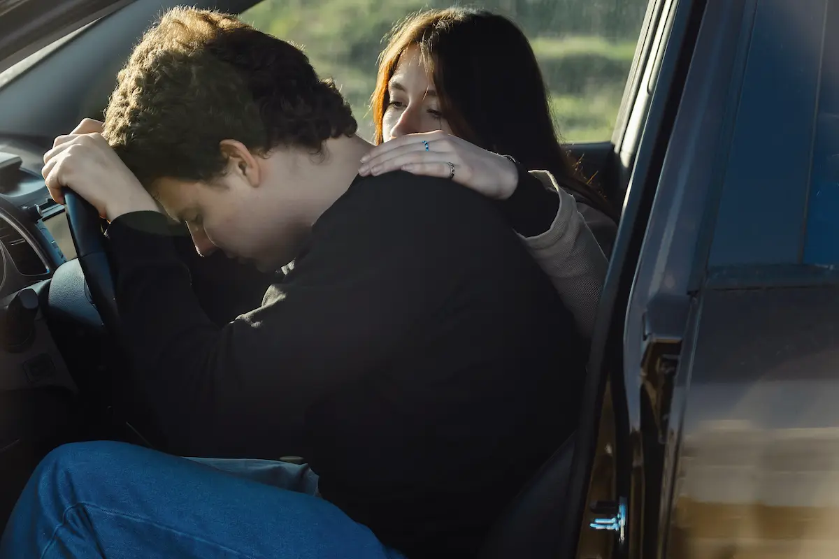 homem vestido de preto sentado em um carro com a cabeça encostada no volante com medo de dirigir e uma mulher ao seu lado o confrota