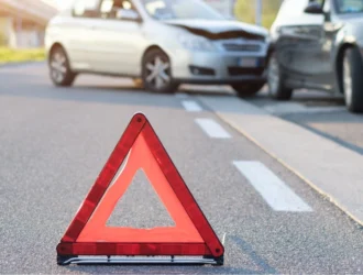 imagem de um triangulo vermelho e alaranjado em uma estrada e ao fundo dois carros acidentados, um preto e um prata