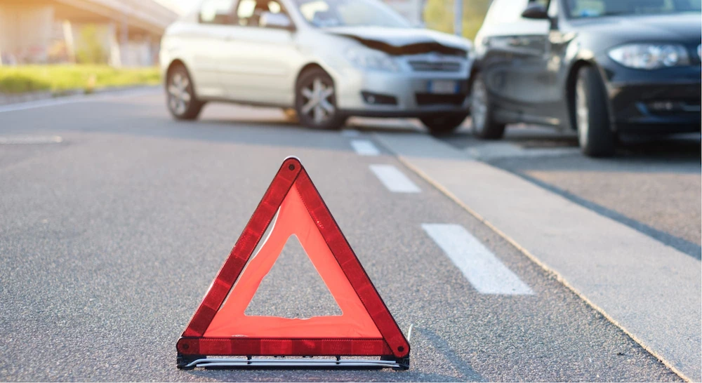 imagem de um triangulo vermelho e alaranjado em uma estrada e ao fundo dois carros acidentados, um preto e um prata