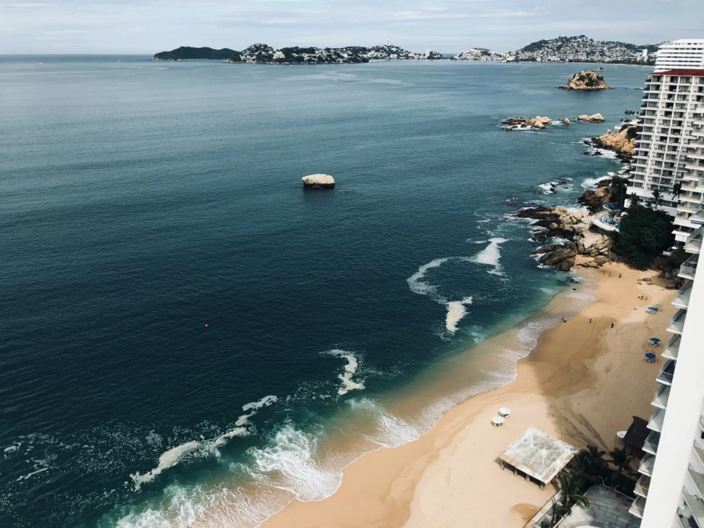 Foto aérea de Acapulco, una de las mejores playas de México, de arena marrón claro, con algunos edificios a la derecha.