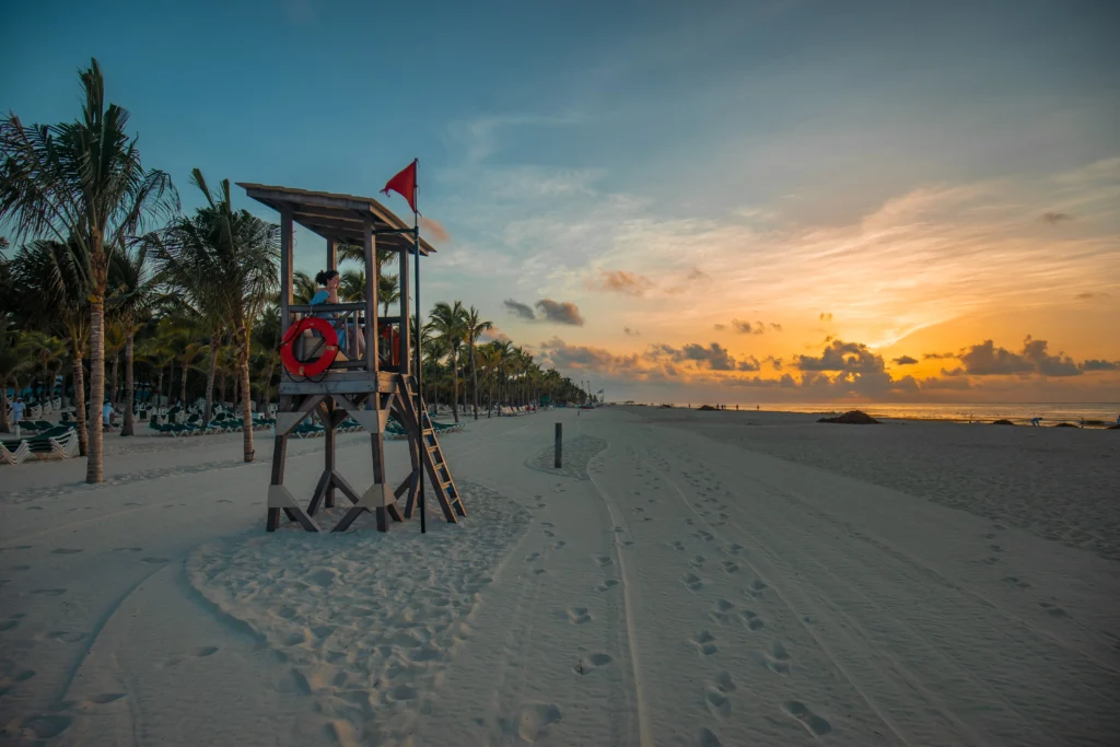 Foto de Playa del Carmen, una de las mejores playas de México, con un cobertizo de salvavidas cerca de la playa, algunas palmeras y el cielo con colores del atardecer.