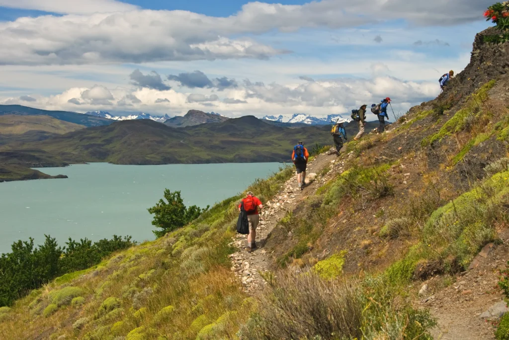 Turistas en un viaje a la Patagonia haciendo senderismo y subiendo una montaña en Torres de Paine, teniendo aguas cristalinas abajo. 