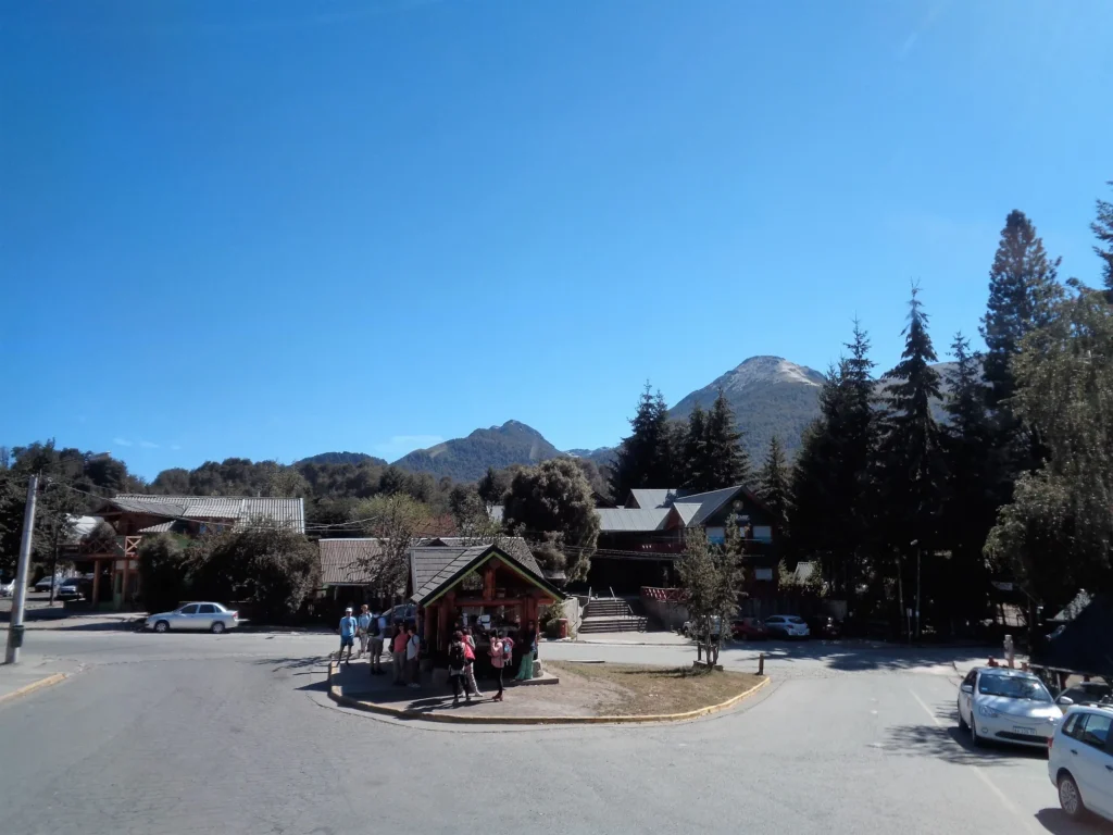Foto de Villa La Angostura, con pequeña casa al centro, pineros a la derecha y la gente alrededor.