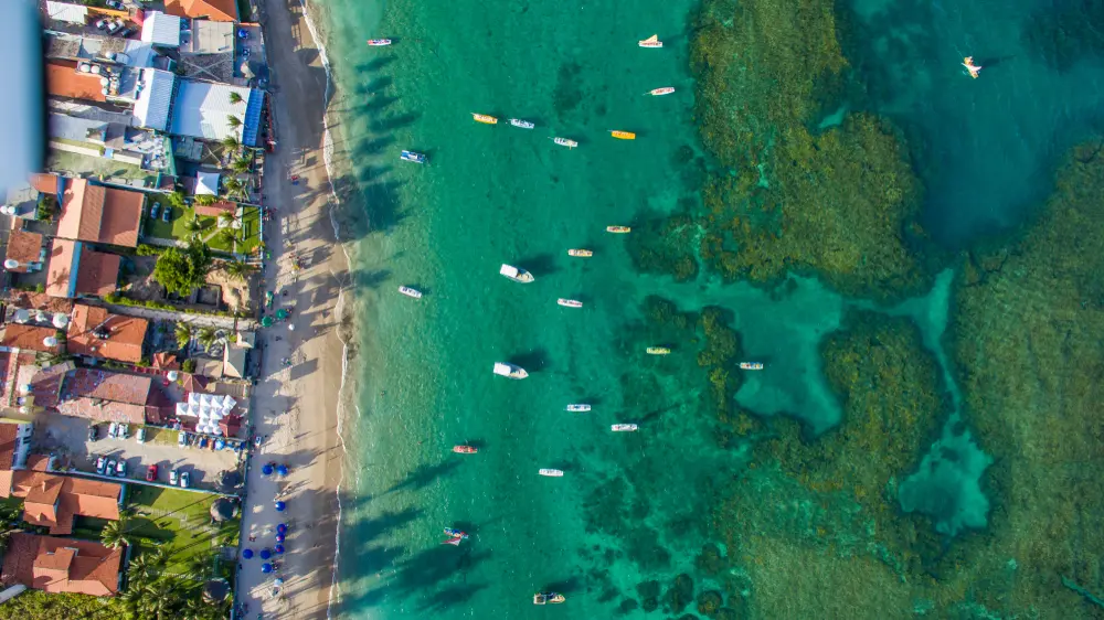 vista aérea do porto de galinhas. a esquerda várias casas típicas a beira mar, e a direita o mar verde esmeralda, com vários barcos e canoas, e abaixo da água cristalina observa-se os recifes