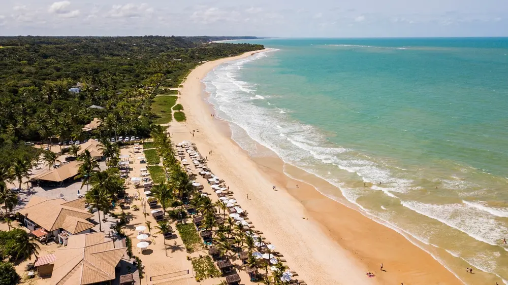 vista aérea de trancoso na Bahia, com muita mata nativa a esquerda, algumas casas abaixo e o mar verde cristalino a direita