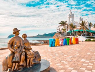estatua de una familia y letras que dicen Mazatlán a la orilla de la playa. Al fondo el edificio Valentinos de mazatlan