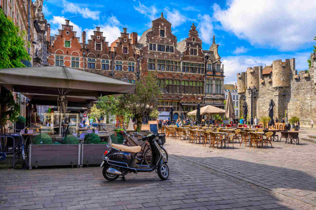 Antiga praça em Gent, Belgica, com mesas ao ar livre e uma moto antiga estacionada.