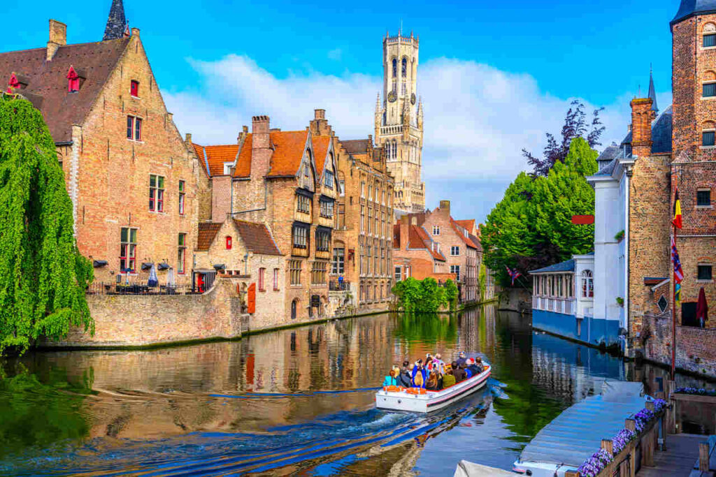 Vista diurna dos canais de Bruges, com um barco de turistas navegando, cercado por construções antigas.