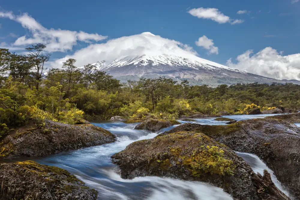 Foto del paisaje de Puerto Varas, con vegetación nativa, una corriente de agua y el volcán Osorno al fundo. 