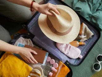pessoa fazendo a mala de acordo com checklist de viagem guardando um chapéu de palha, cosméticos e passagens aéreas