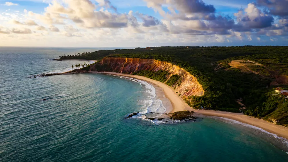 Praia de João Pessoa, na Paraíba, onde o mar é vasto e azul, com uma faixa de areia pequena, um grande relevo na pedra e uma área verde.