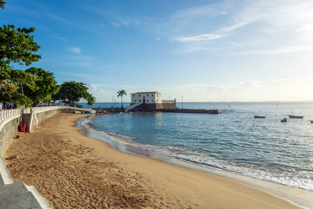 Foto de uma praia de Salvador, capital da Bahia. É possível ver o céu bem azul e o mar calmo. Ao fundo há uma construção isolada.