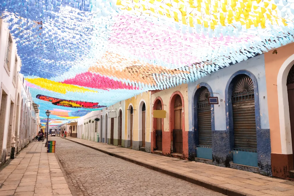 Foto de uma das ruas do centro histórico de São Luís, capital do Maranhão. Há bandeirinhas de São João suspensa ao alto, parecendo um teto que liga os dois lados da rua.