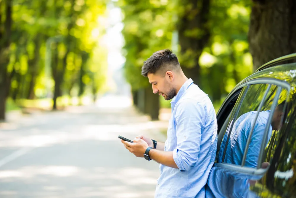 Homem mexendo no celular, encostado na porta do carro. Ao fundo, temos uma floresta.