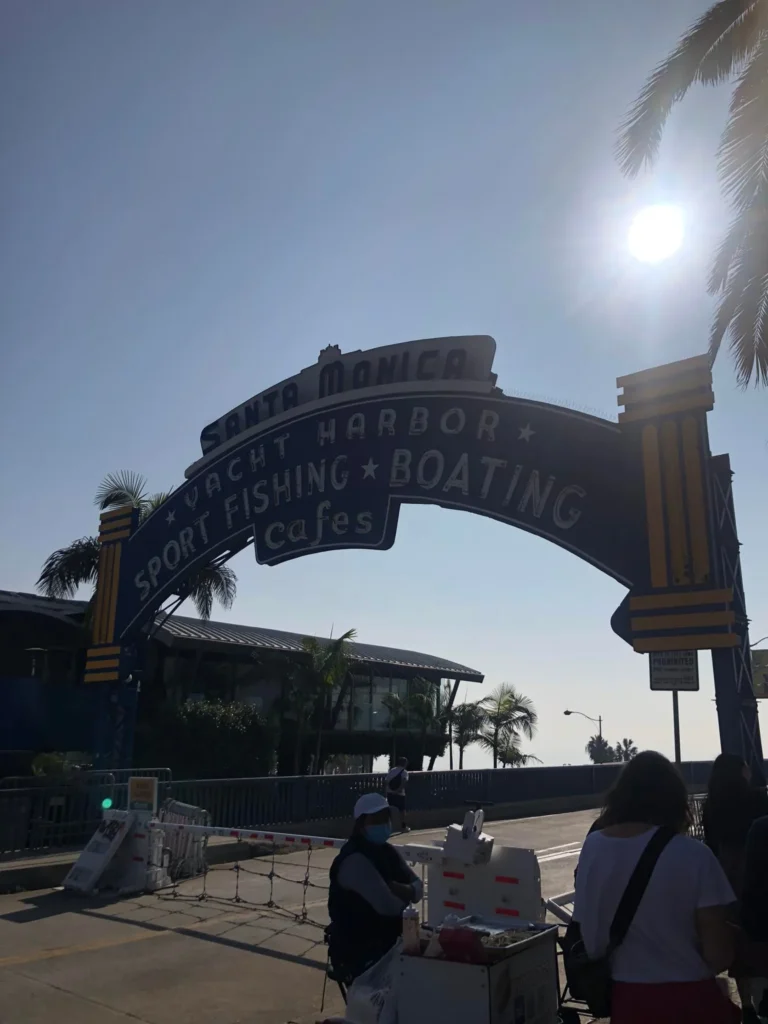Foto do letreiro que indica a entrada do Píer de Santa Mônica, onde é possível ver 2 pessoas em frente ao letreiro. O dia está bem claro.