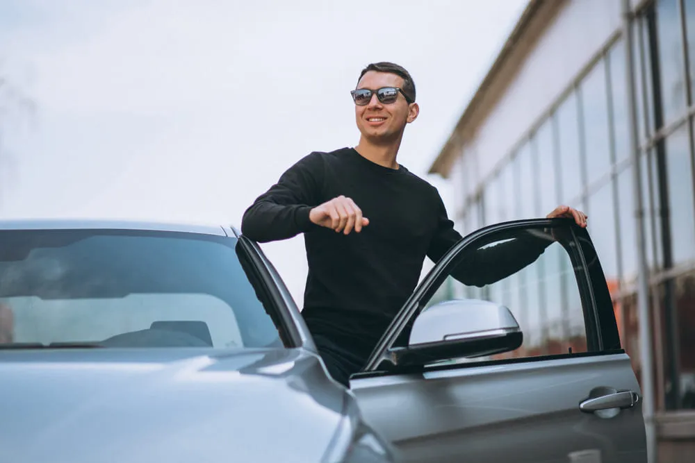 Homem sorrindo ao lado do seu carro. O dia está nublado. 