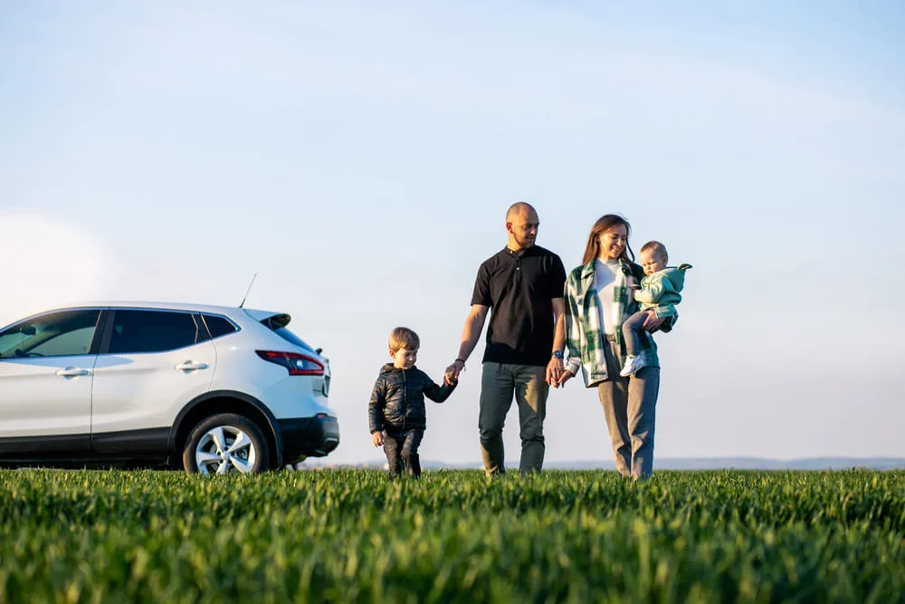 Foto de um pai, uma mãe e dois filhos perto do carro, um SUV na cor branca. A família está de mãos dadas e felizes em estar viajando. O solo é repleto de grama. verde.