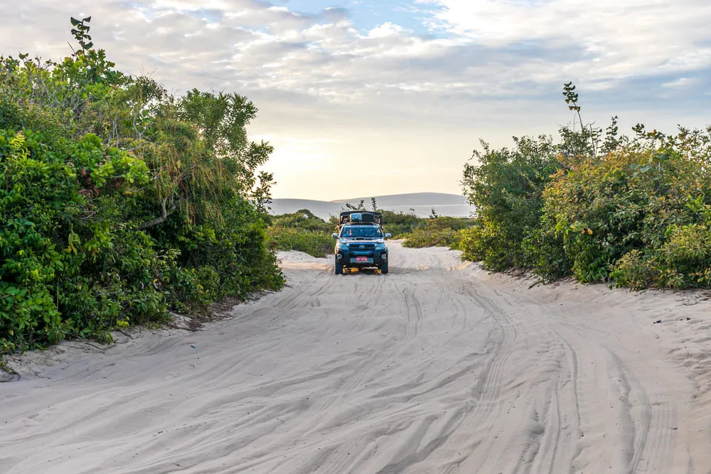 Foto de um carro SUV 4x4 com pessoas passeando por Jericoacoara, região Nordeste do Brasil. A estrada é de areia e há vegetação verde em volta.