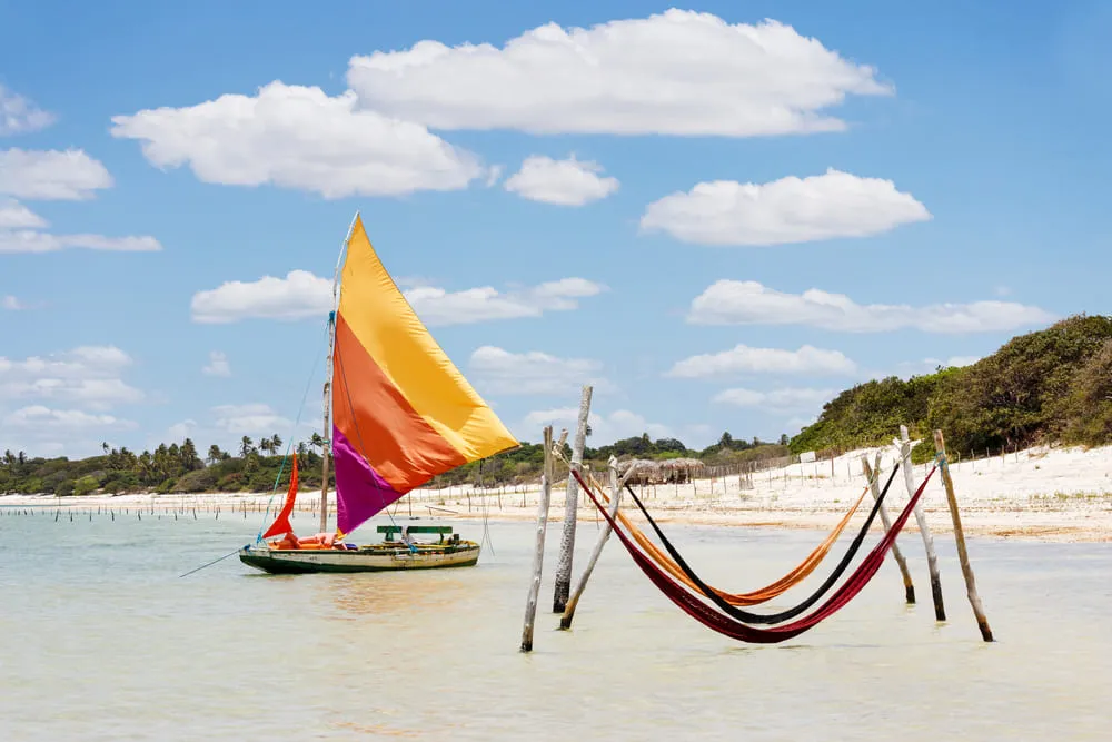 Foto da Lagoa do Paraíso em Jericoacoara, região Nordeste do Brasil. Há 2 redes na parte rasa do mar, próprias para o descanso. Além disso, há um pequeno barco colorido no mar. O céu está azul e o mar calmo.