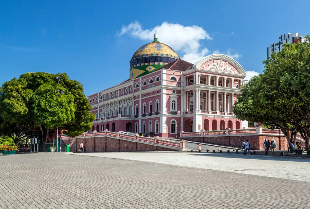 Foto de um dos monumentos mais importantes de Manaus, Amazonas, o Teatro Amazonas. Ele é rosa e branco. O céu está azul e algumas pessoas circulam em volta do monumento.