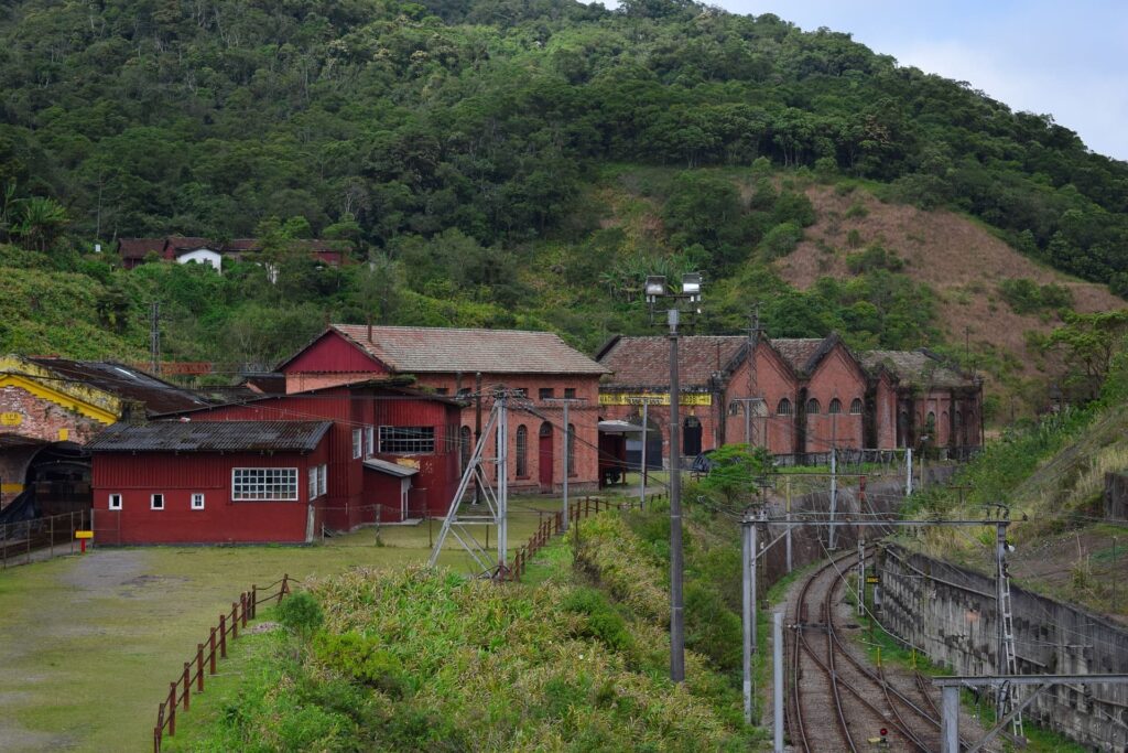 Foto das casas vermelhas da cidade de Paranapiacaba, São Paulo. A vegetação ao redor é verde e há um morro ao fundo. A linha de trem fica ao lado da vila.