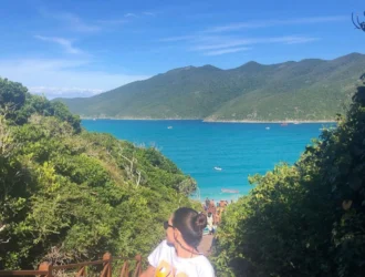 Foto da praia de Portal do Atalaia em Arraial do Cabo. A foto foi tirada a partir da vista do alto da escadaria. É possível ver o mar bem azul, quase verde, o céu bem claro e pessoas subindo e descendo a escada. Uma mulher está bem ao centro da foto posando para a mesma.
