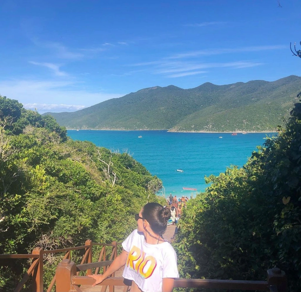 Foto da praia de Portal do Atalaia em Arraial do Cabo. A foto foi tirada a partir da vista do alto da escadaria. É possível ver o mar bem azul, quase verde, o céu bem claro e pessoas subindo e descendo a escada. Uma mulher está bem ao centro da foto posando para a mesma.