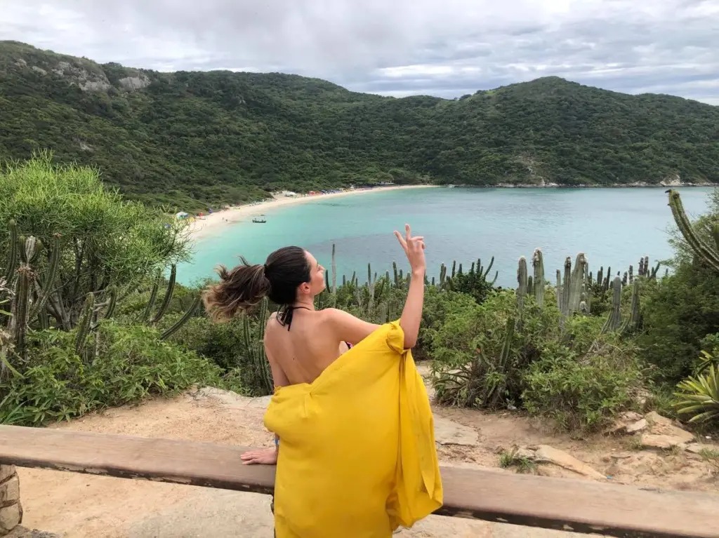 Foto do alto do morro onde é possível observar a Praia do Forno em Arraial do Cabo, no Rio de Janeiro. A vista mostra o mar bem verde e a vegetação em volta. O dia está nublado. Ao centro está uma mulher com uma camisa amarela posando para foto.