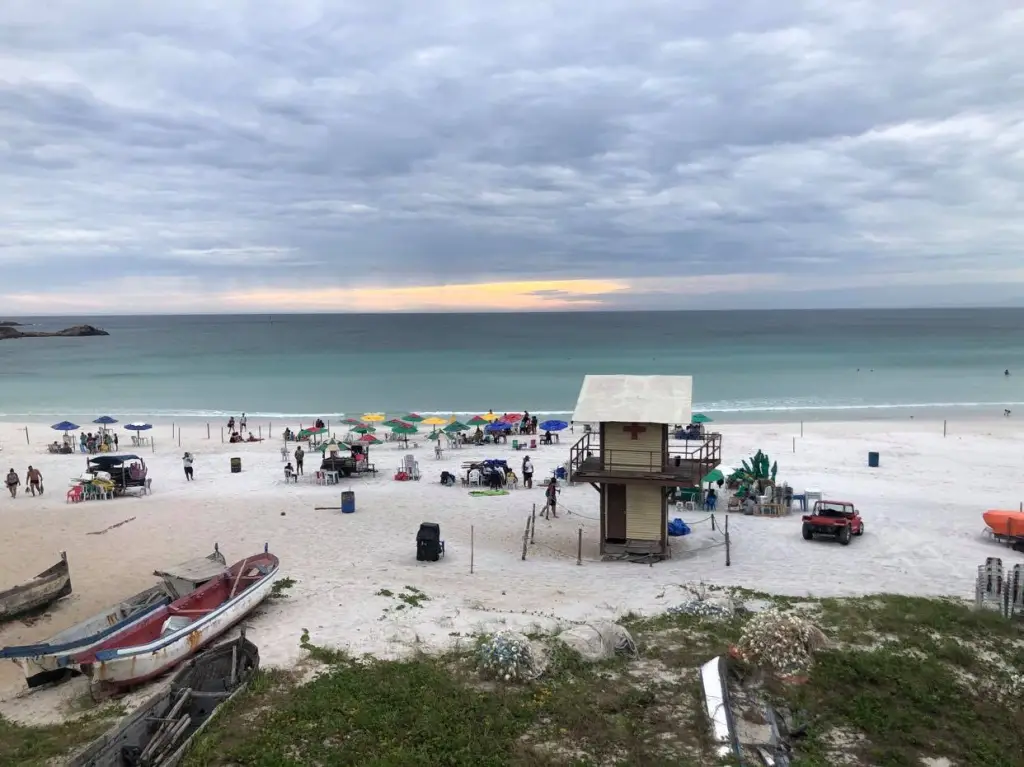 Foto da Praia Grande em Arraial do Cabo. O dia está bastante nebuloso, mas é possível ver um resto de pôr do sol ao fundo, deixando uma parte do céu amarelo. A praia conta com pessoas na areia e algumas no mar. Tem 4 barcos na areia e uma casa de salva vidas ao centro.