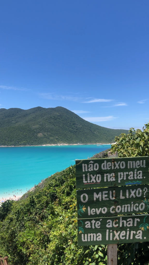 Foto da praia de Portal do Atalaia em Arraial do Cabo. A foto foi tirada a partir da vista do alto da escadaria. É possível ver o mar bem azul, quase verde, o céu bem claro e as placas que dizem "Não deixo meu lixo na praia" "O meu lixo? Levo comigo" "Até achar lixeira".