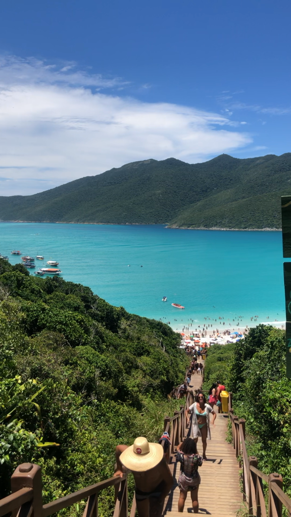 Foto da praia de Portal do Atalaia em Arraial do Cabo. A foto foi tirada a partir da vista do alto da escadaria. É possível ver o mar bem azul, quase verde, o céu bem claro e pessoas subindo e descendo a escada.
