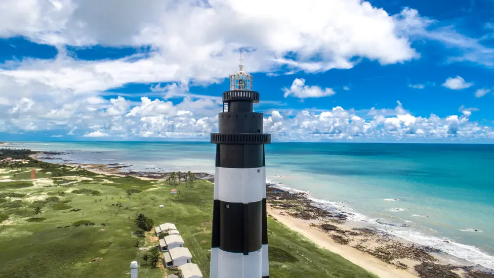 imagem de um farol pintado com faixas horizontais em preto e branco. ao fundo a esquerda, vegetação nativa, a esquerda o mar azul