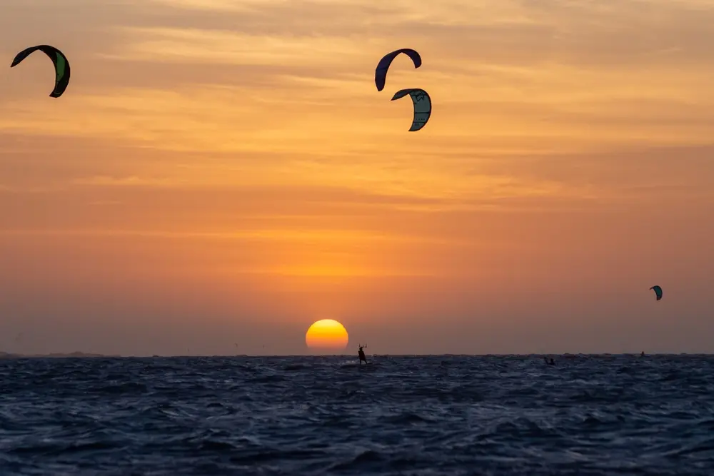 pôr do sol visto do mar azul escuro, com o sol tocando o horizonte e o céu na cor laranja enquanto quatro kitesurfs  estão praticando
