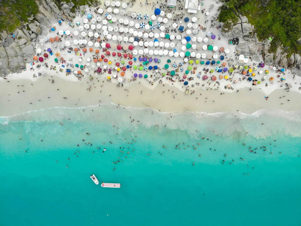 Foto aérea da praia de Portal de Atalaia em Arraial do Cabo no Rio de Janeiro. O mar é bem azul com a vegetação verde em volta. É possível ver todos os guarda-sóis na areia, com muitas pessoas tomando sol e banho de mar.