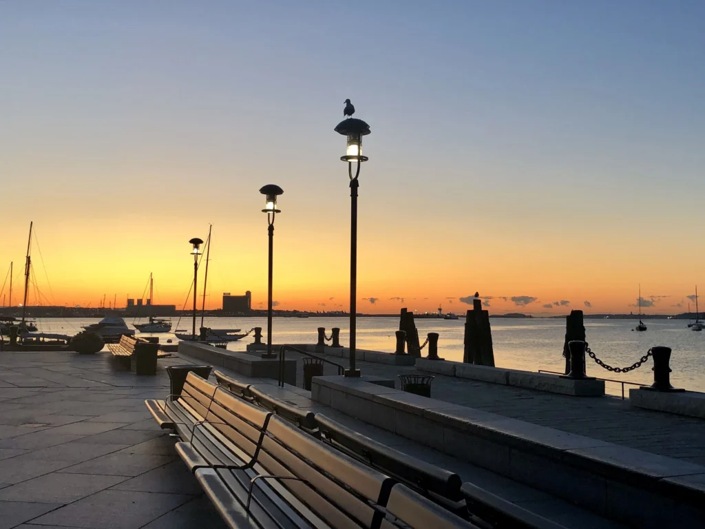 Foto do entardecer na cidade de Boston em Massachusetts. É possível ver o píer, o mar e alguns assentos com lustres ao lado.