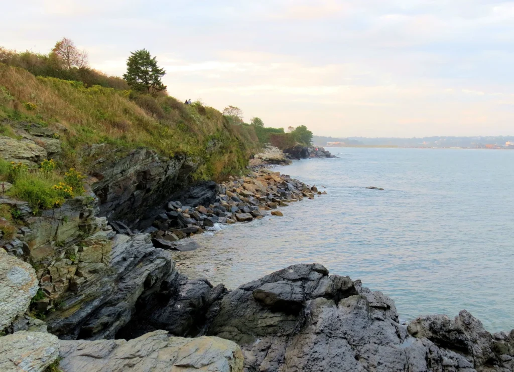Foto da costa de Newport, em Rhode Island. É possível ver o mar batendo nas pedras e o céu está nublado.