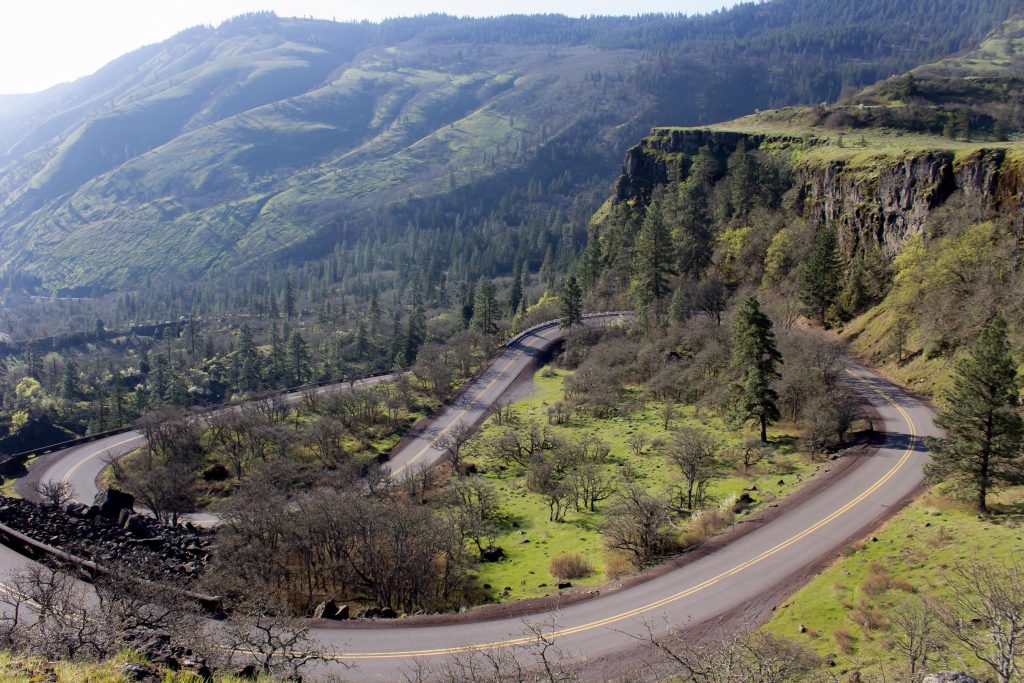 Foto do alto da Historic Columbia River Highway, uma das estradas consideradas perigosas por conta de suas curvas sinuosas. Ao redor há muitas montanhas e rochas imponentes. A estrada está vazia.