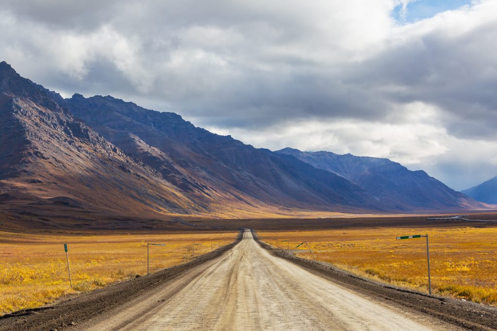 Foto da James Dalton Highway, uma estrada longa e de terra. Ao fundo é possível ver diversas montanhas de terra. O solo é bem árido e a vegetação parece bem seca.