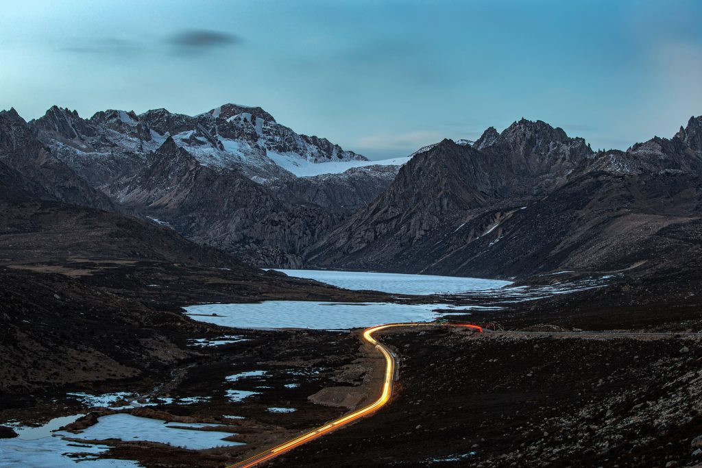 Foto de uma estrada em meio às rochas nevadas em Sichuan Tibet. Está anoitecendo e é possível ver as luzes dos carros na estrada.