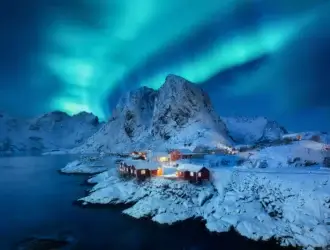 Foto incrível de uma aurora boreal acontecendo na Noruega. Na foto, ainda aparecem os montes todos nevados e as casas, também nevadas, abaixo. Um lago permeia a região.