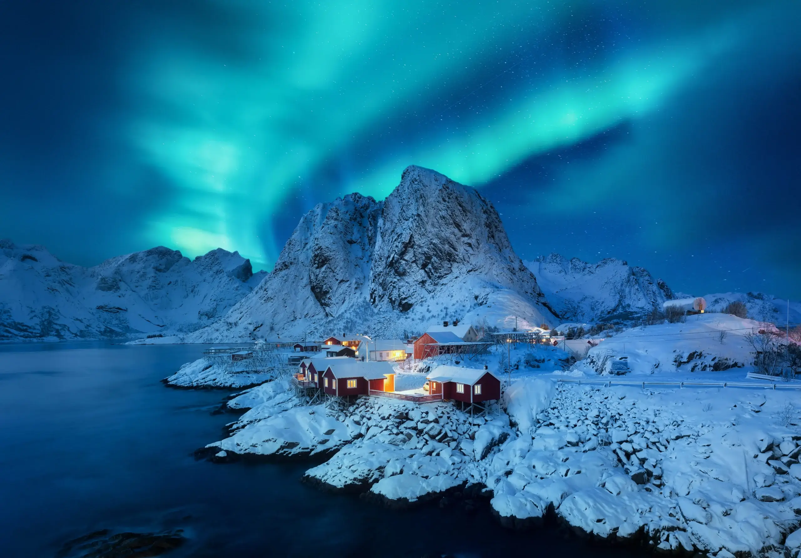 Foto incrível de uma aurora boreal acontecendo na Noruega. Na foto, ainda aparecem os montes todos nevados e as casas, também nevadas, abaixo. Um lago permeia a região.