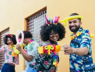 Foto de quatro pessoas comemorando o Carnaval. As roupas são coloridas e eles carregam elementos da festa: bexiga colorida, máscaras coloridas e acessórios para o cabelo. Todos sorriem para foto.