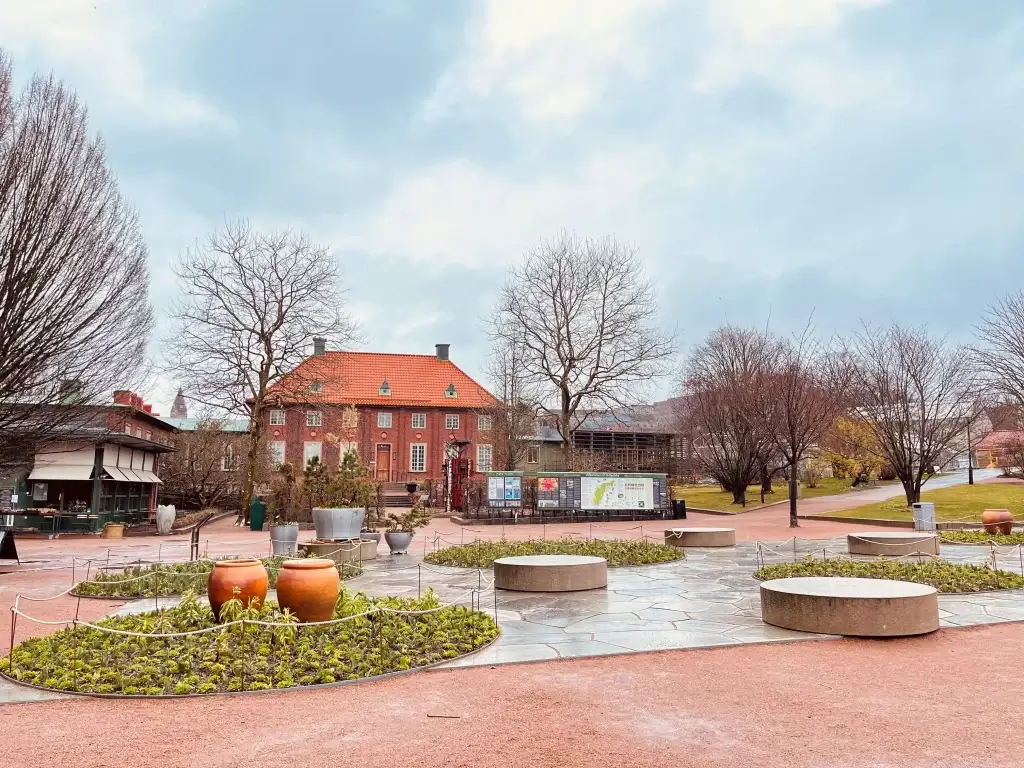 Foto de uma vila de Gotemburgo, onde é possível ver casas ao fundo com uma pequena praça em frente. Há vegetação verde e também seca, com o céu azul ao fundo.