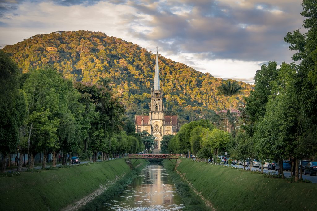 Foto de Petrópolis, cidade do Rio de Janeiro, com a catedral ao fundo e o monte repleto de vegetação. Há um riacho em frente e o céu está nublado.