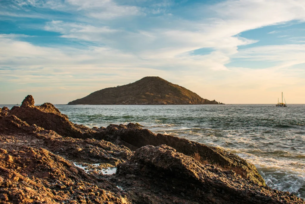 Foto del paisaje de uno de los lugares para visitar en Mazatlán, con el cielo azul, algunas piedras, el mar y una montaña al fondo.