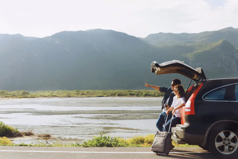 Pareja con maleta en manos y con el maletero de un auto abierto. El hombre apunta lejos y detrás de la pareja hay un lago y montañas.
