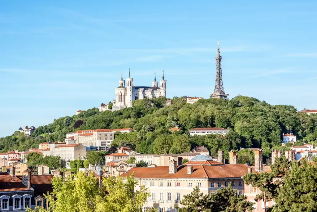 Foto de um morro na cidade de Lyon, na França. É possível ver um castelo no alto em meio a vegetação verde e uma réplica da Torre Eiffel. É possível ver casas francesas entre as árvores. O céu está azul.