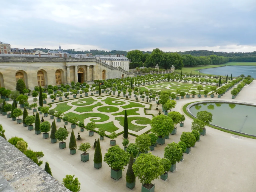 Foto do jardim do Palácio de Versalhes. É possível ver as infinitas flores bem verdes e com um paisagismo incrível. Tem um lago ao fundo e o palácio ao lado.