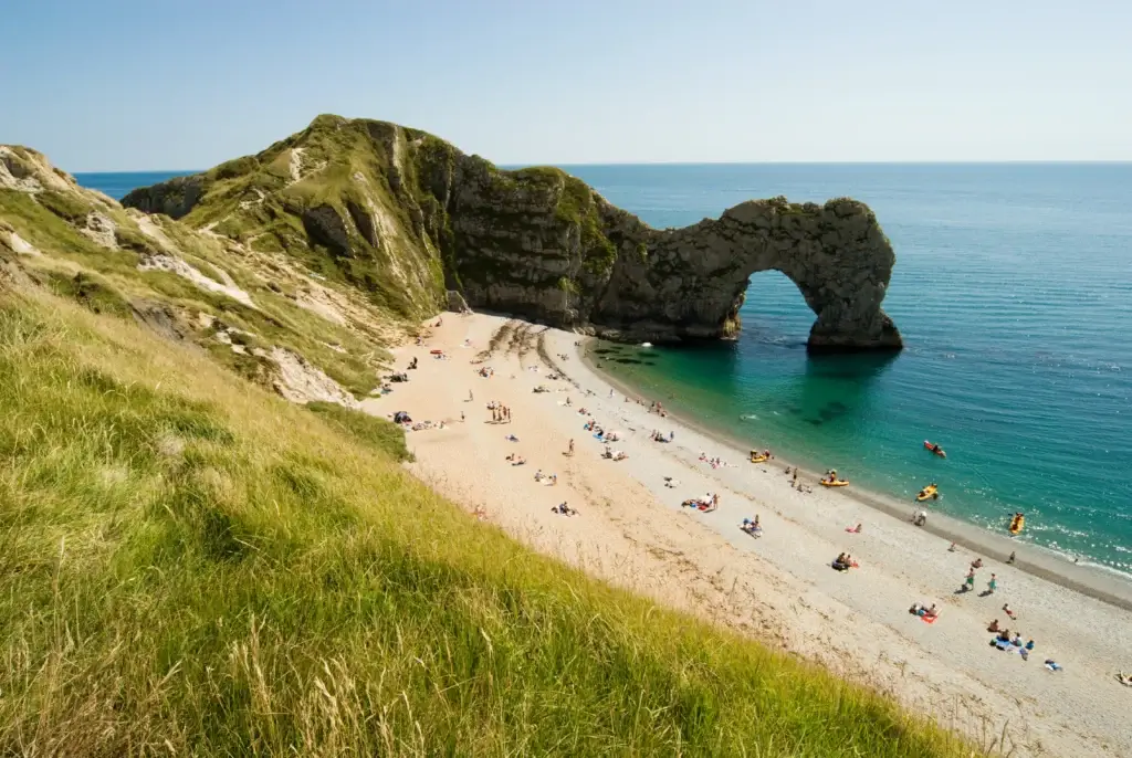 Image of a rocky outcrop on the Dorset coast, with the English Channel in the background. The beach is visible below, with people sunbathing on the sand.